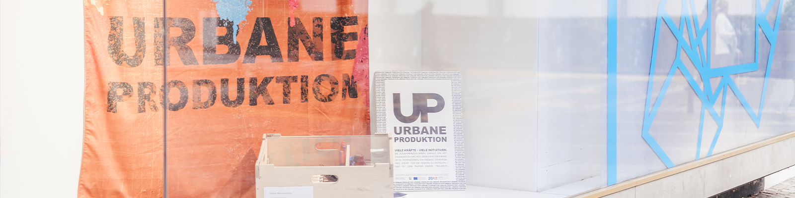 Zusehen ist ein Schaufenster der gläsernen werkstatt, in welchem ein orangenes Tuch mit der Aufschrift "Urbane Produktion" hängt. Davor steht eine helle Holzkiste und ein weißes Schild mit "UP Urbane Produktion" und kleinerem Text. 